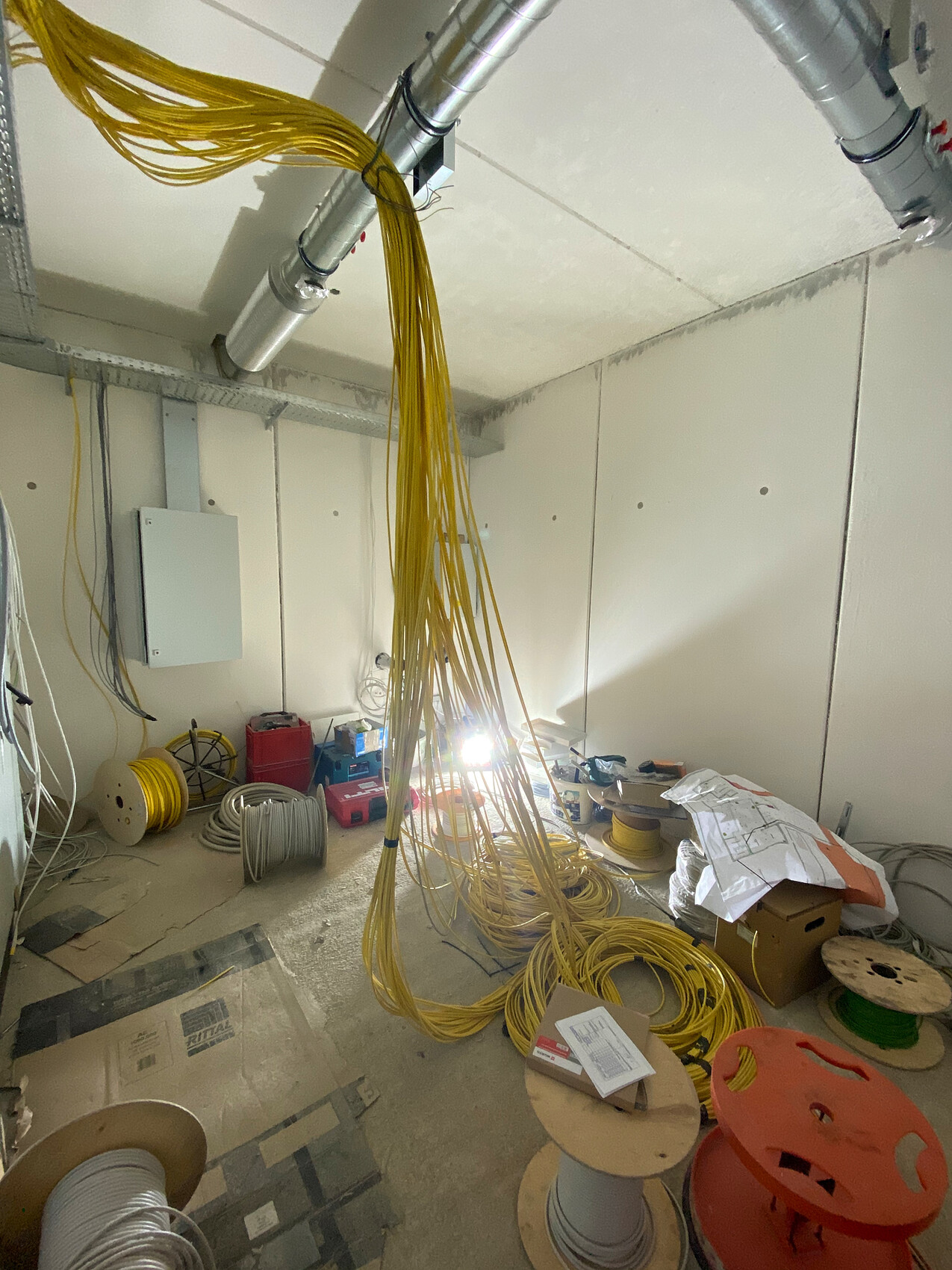 Viele gelbe Kabelstränge hängen von der Decke; Kabelrollen und Werkzeuge liegen im Technikraum.
