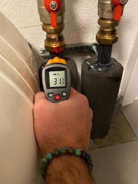 Hand hält digitales Messgerät vor Heizungsrohr-Verbindung; Anzeige 31,5 °C.