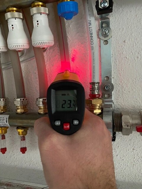 Hand hält digitales Temperaturmessgerät an Heizungsrohrverteilung; Display zeigt 23,4°C