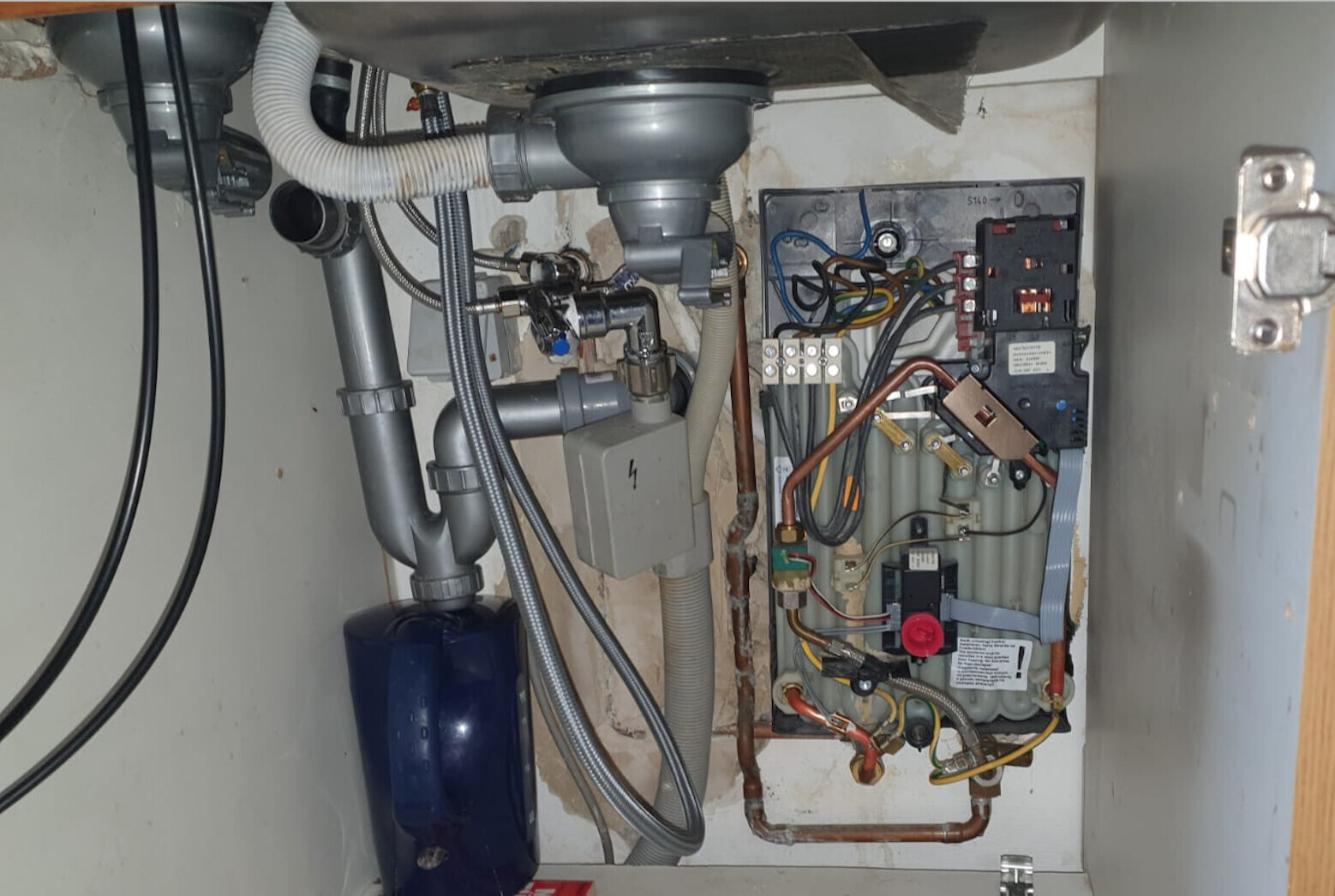 Unter dem Spülbecken: Rohre, P-Trap, Schläuche und Heizungs-/Wasseranlage mit Steuerung.