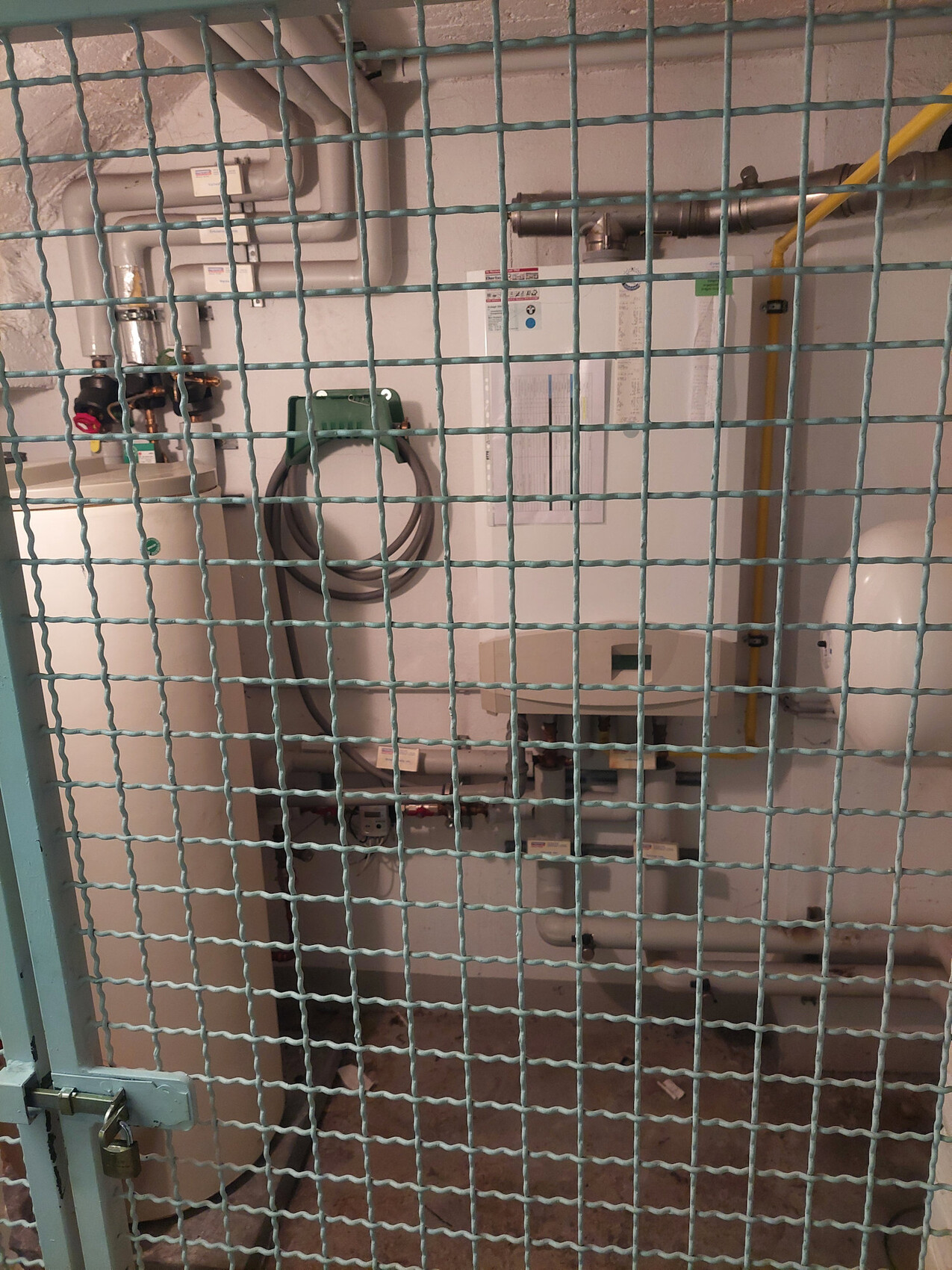 Blick durch grünes Metallgitter auf eine Heizungsanlage/Boiler mit Rohren im Technikraum