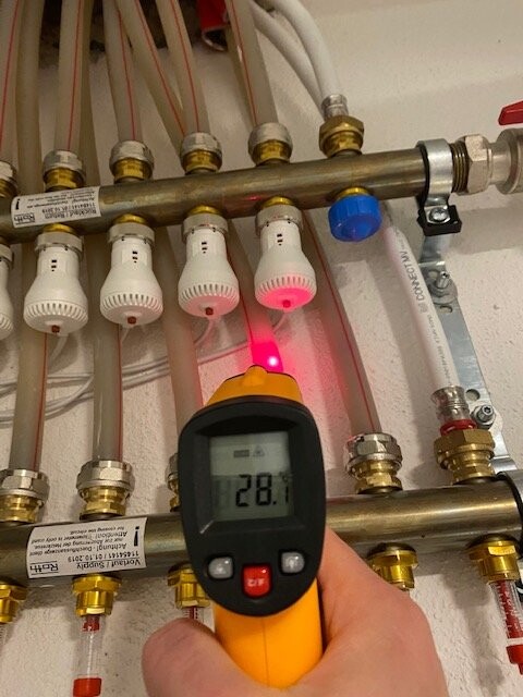Hand hält Infrarot-Thermometer an Heizungsrohr mit TRV-Ventilen, roter Laser sichtbar.