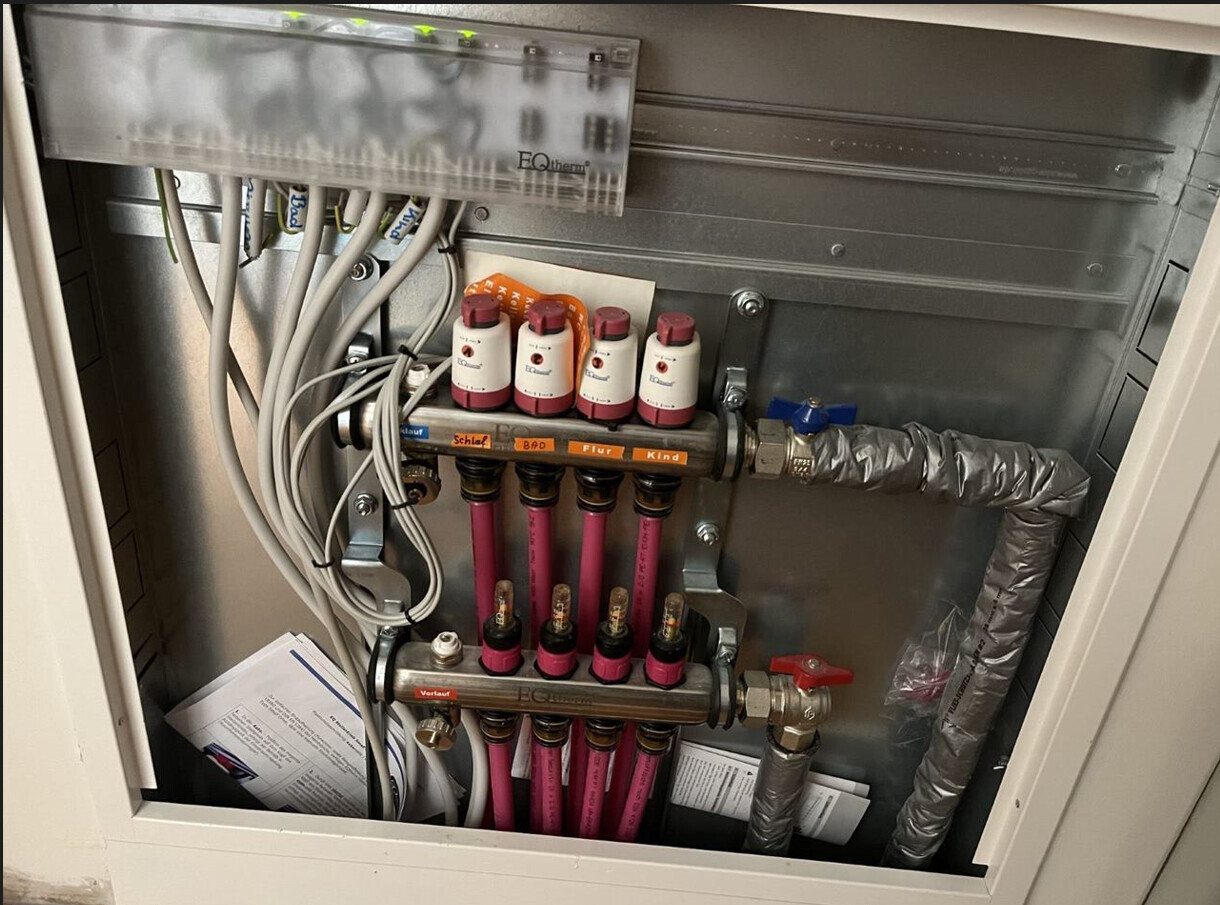 Heizungsverteiler mit pinken Rohren, Ventilen, Kabeln und Isolierung in einer Metallverkleidung.