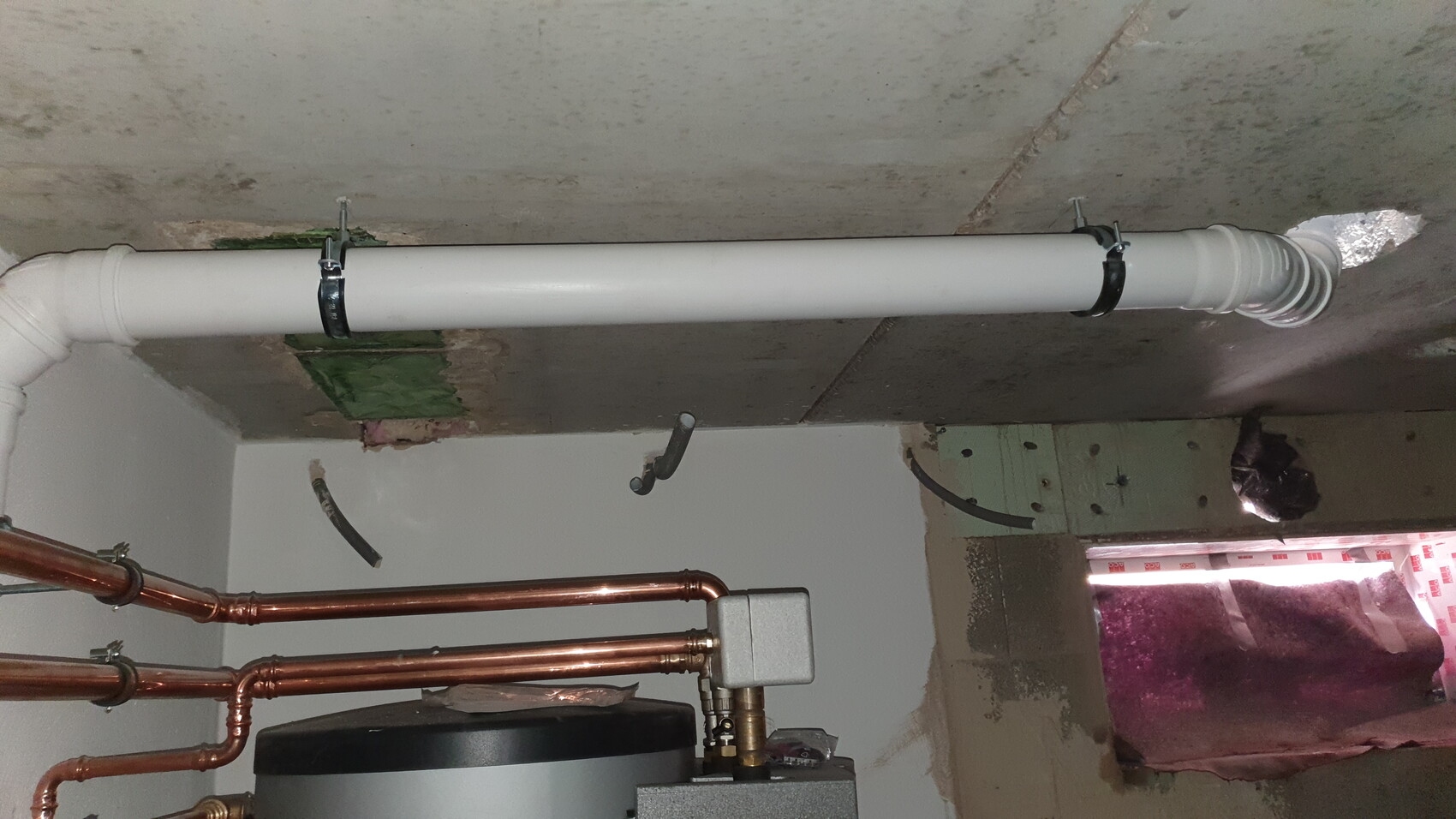 Weißes PVC-Abflussrohr an der Decke mit Klemmen, daneben Kupferrohre und Heizgerät.