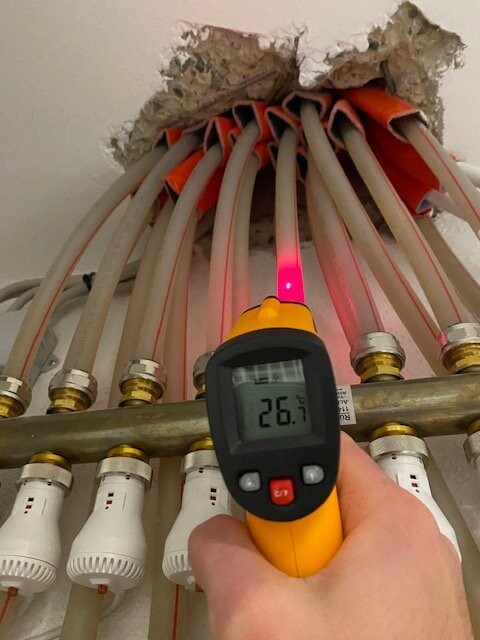 Hand hält Infrarot-Thermometer vor rotes Rohrleitungsbündel durch Loch in Decke; Anzeige 26,7 °C