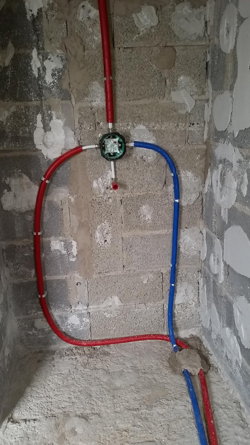 Rote und blaue Wasserleitungen führen zu einem runden grünen Verteiler an einer Rohbauwand.