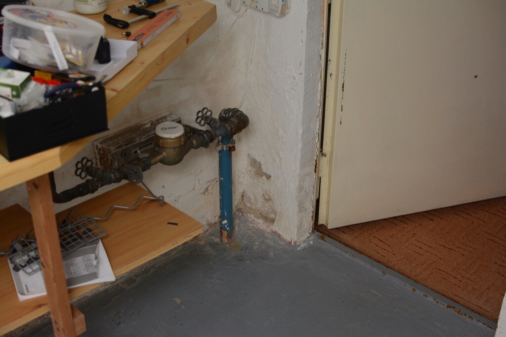 Offene Wasserrohre und Wasserzähler an der Wand neben einer Holzwerkbank mit Werkzeugen.