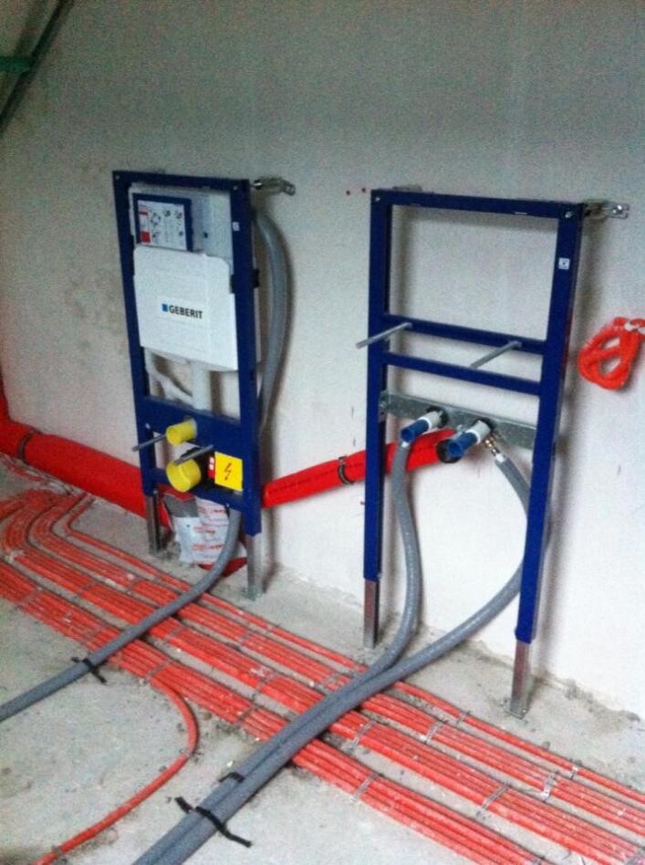 Zwei blaue Metallrahmen mit Sanitär- und Heizmodulen; rote Bodenrohre und graue Leitungen.