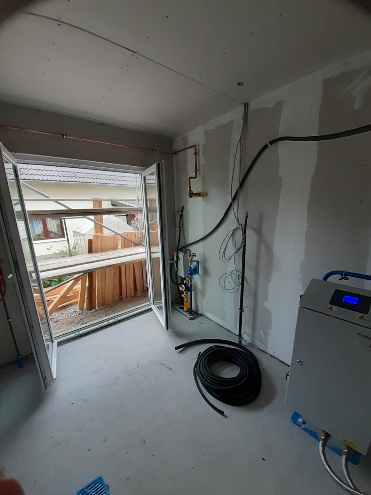 Baustellenraum: offener Balkon, sichtbare Rohre, Kabel, Schlauchrolle, Heizgerät rechts.