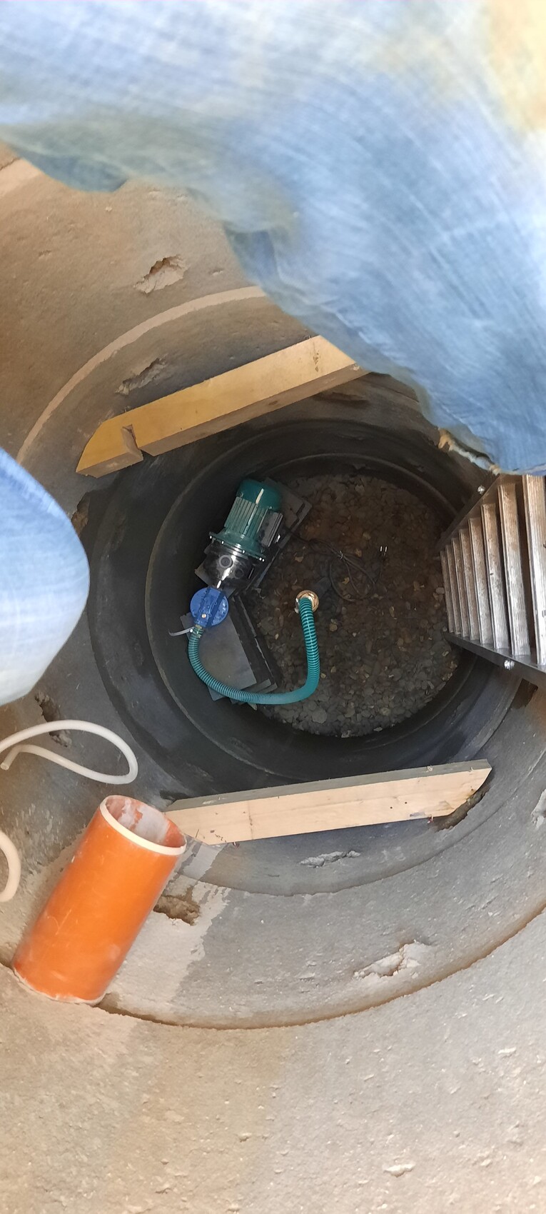 Runder Schacht mit grüner Pumpe, blauem Schlauch und Leiter im Baugrubenbereich
