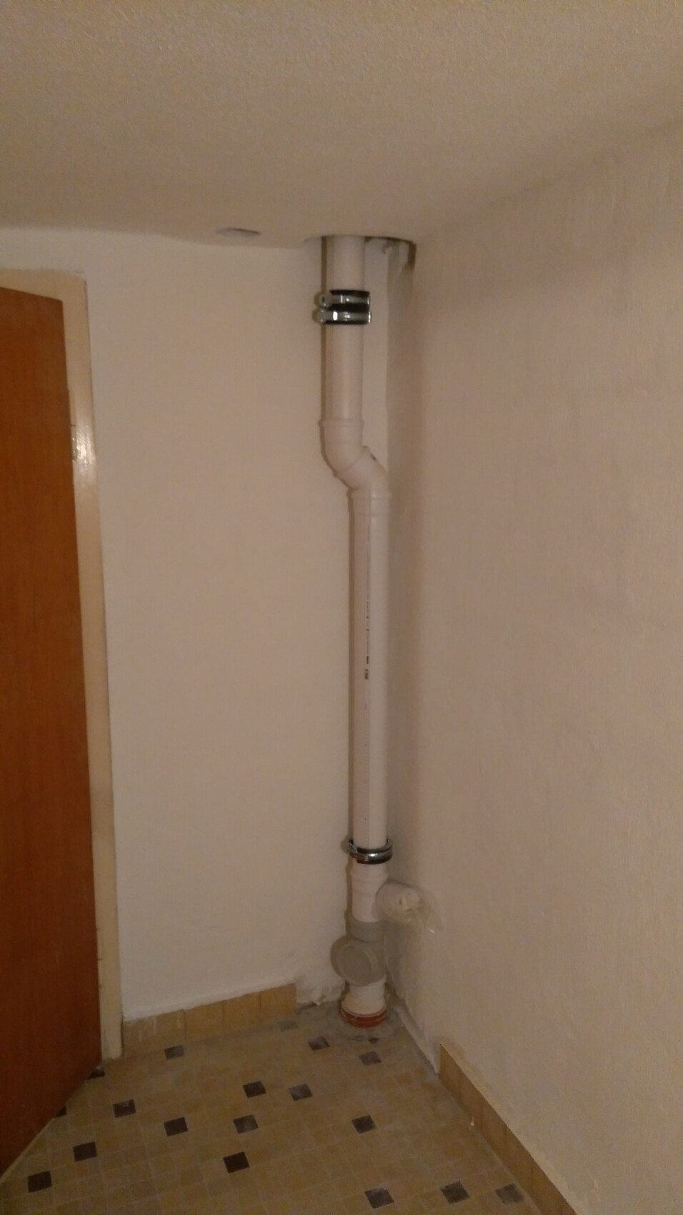 Weiße PVC-Abflussleitung mit Bögen an der Wand in der Ecke, mit Metallklemmen und Bodenanschluss.
