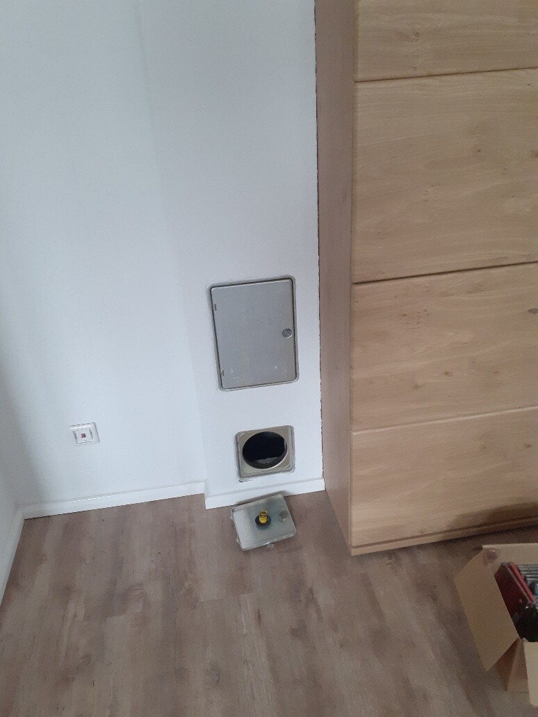 Eckwand eines Zimmers mit runder Öffnung unten im Wandbereich und Metallabdeckung daneben; Holzboden.