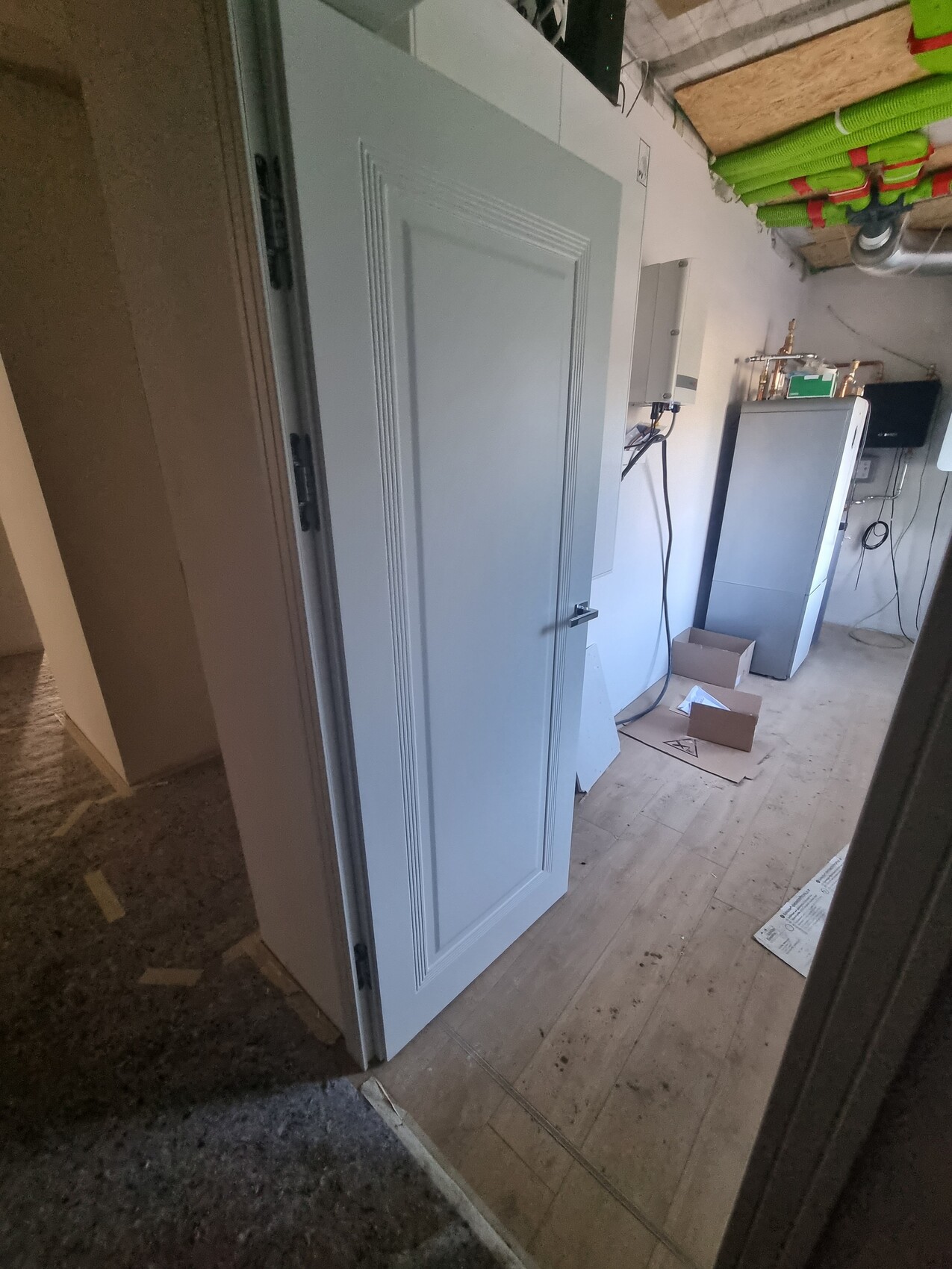 Technikraum mit offener Tür, größer grauer Schrank, Boiler und Kartons auf Holzboden.