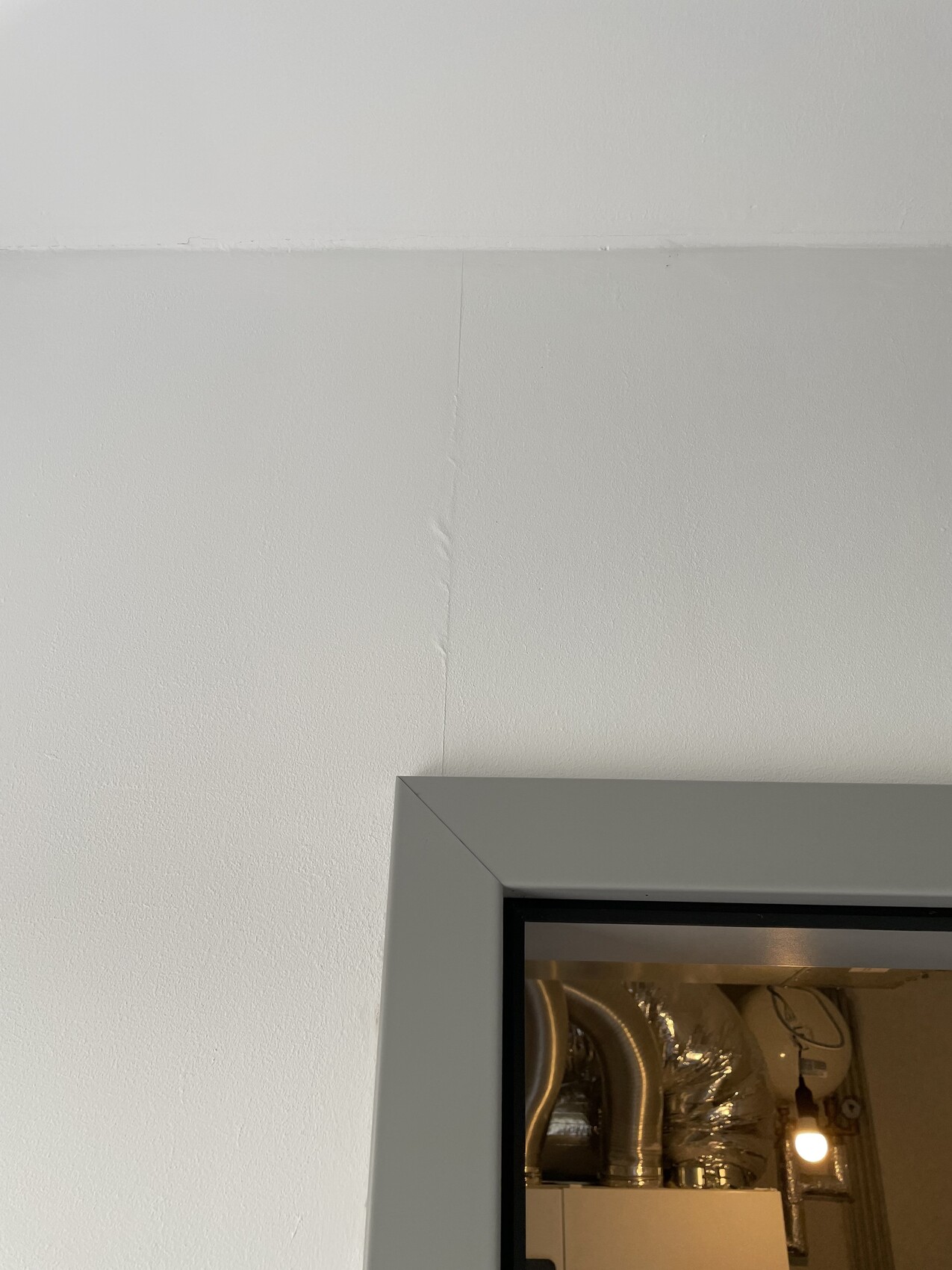 Türrahmen aus grauem Metall vor weißer Wand; dahinter Lüftungsrohre und Lampenlicht.