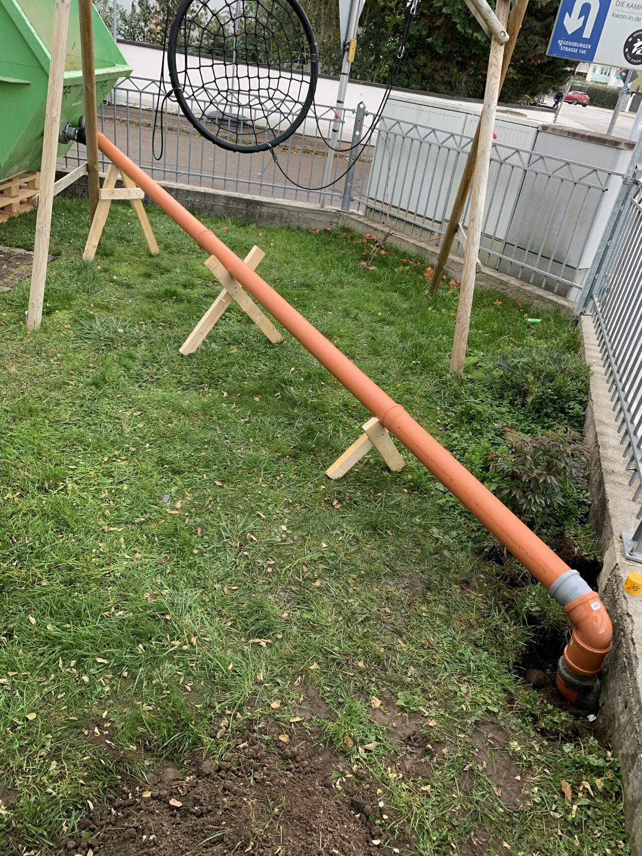 Diagonal verlegtes orangenes Rohr, auf Holzstützen gestützt, führt zu Bodeneinlass am Spielplatz