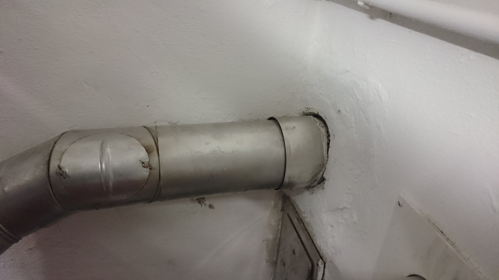 Metallisches Lüftungsrohr mit Bögen durch eine Wandöffnung, weißer Putz darum herum.