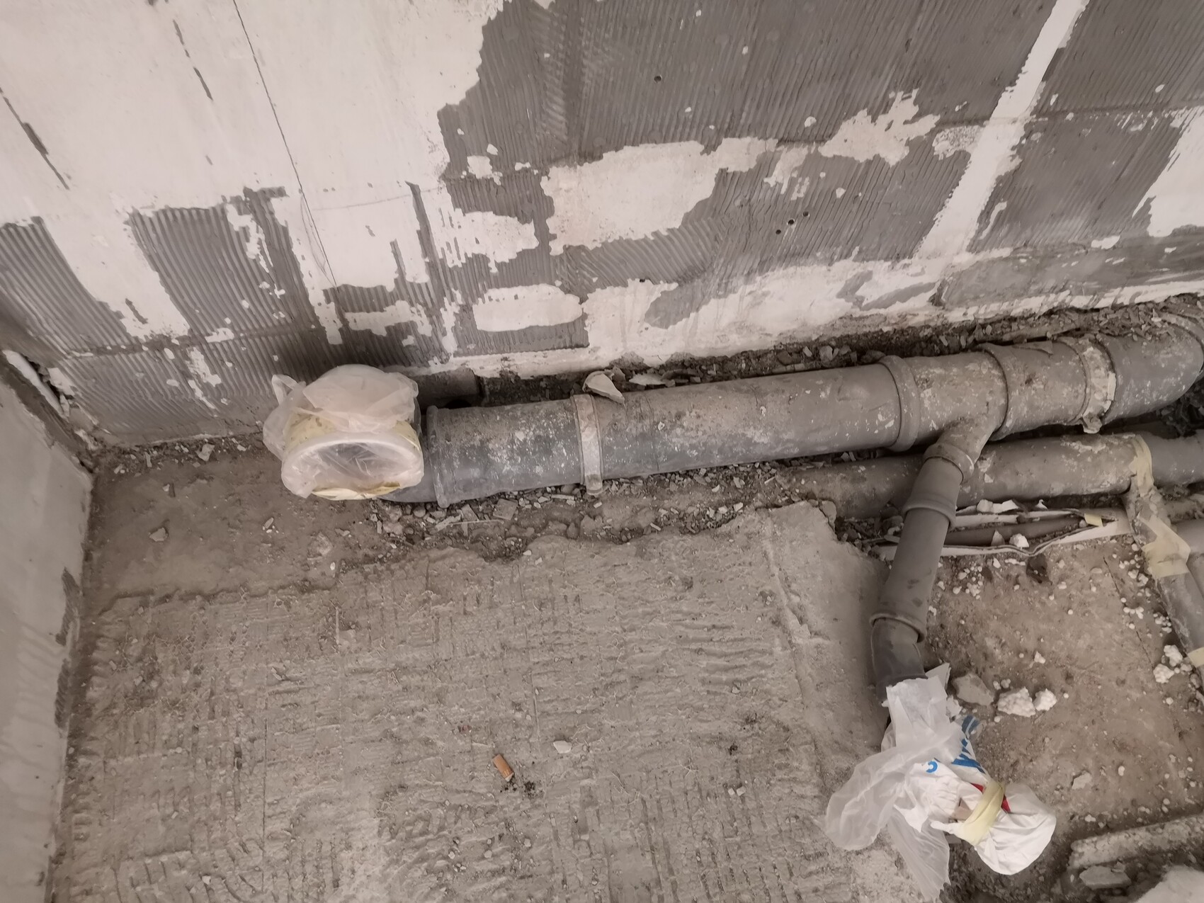 Verlegte Rohre entlang einer rauen Baustellenwand; Plastikabfälle und Müll liegen am Boden.