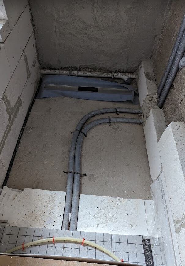 Kleine Baugrube mit verlegten grauen Rohren auf dem Betonboden; hinten Abflussdeckel sichtbar.