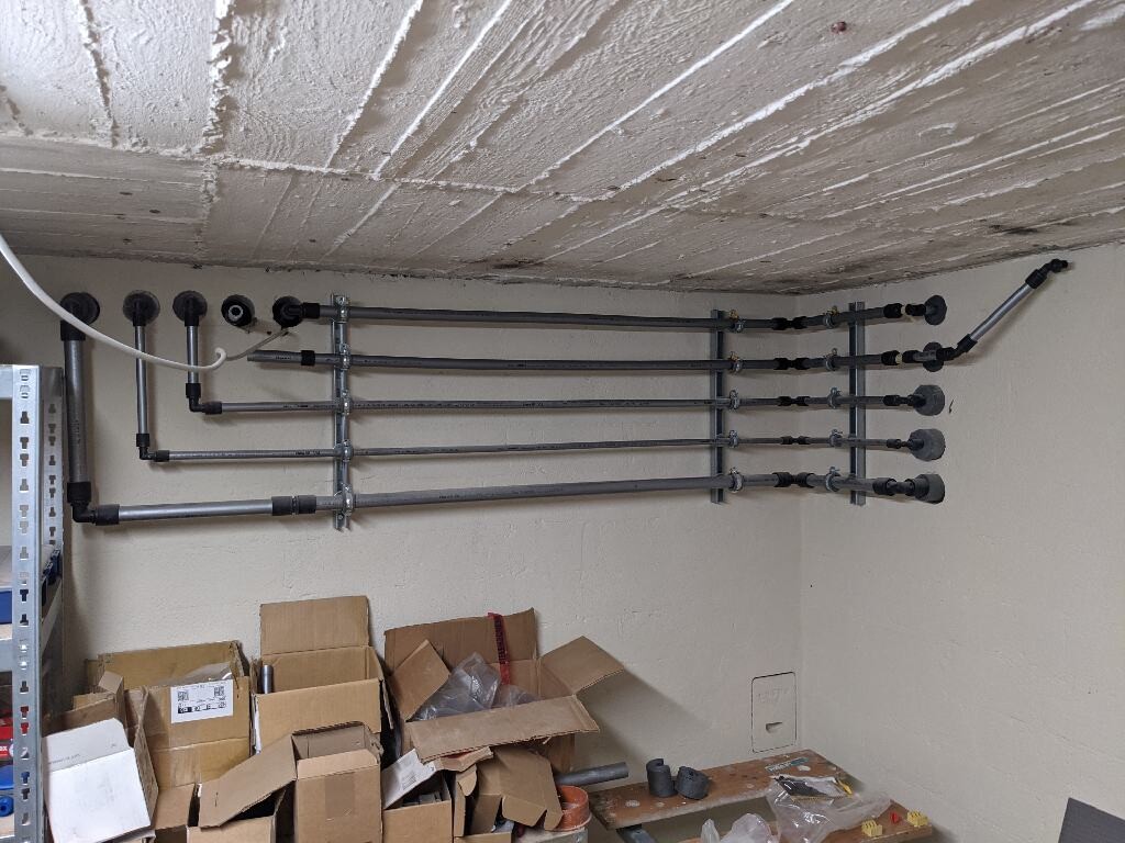 Mehrere Metallrohre verlaufen waagerecht an Wand und Decke in einem Kellerraum; Kartons unten.