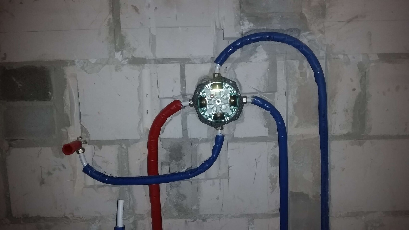 Runde Metallverbindung mit vier Anschlüssen; rotes und blaues Rohr im Keller.