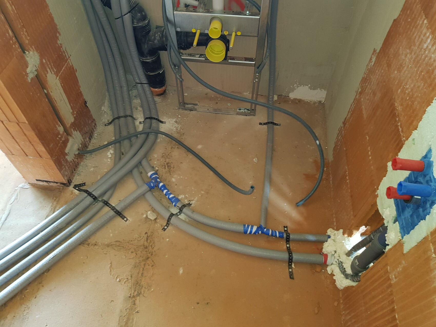 Baustellensituation: Mehrere graue Rohre, Schläuche und Kabel liegen am Boden vor einer Wand.