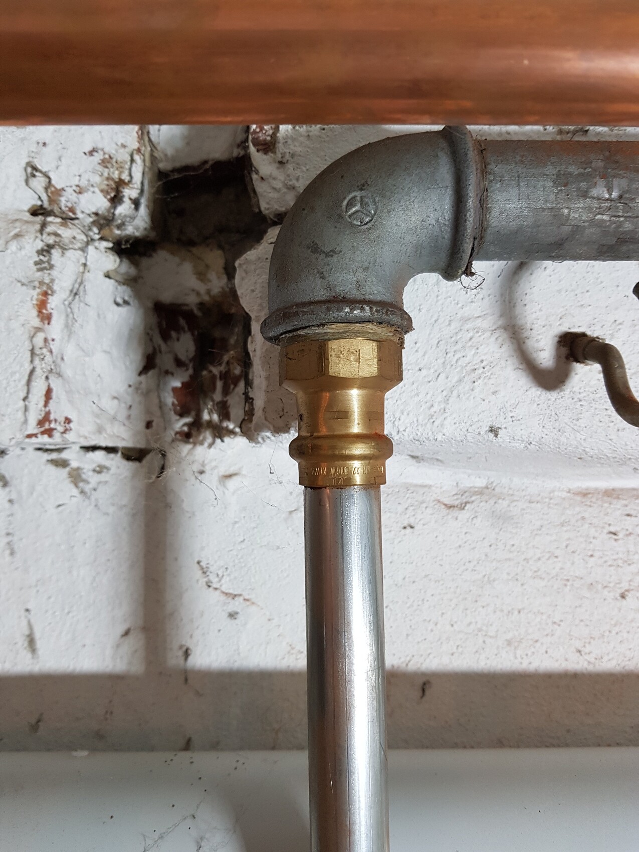 Wasserleitung: 90°-Bogen aus Metall mit Messing-Kompressionsmuffe, vertikales Rohr darunter.