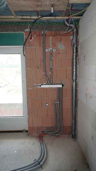 Rohbau mit sichtbaren Wasser- und Elektroleitungen an Ziegelwand, Fenster links.