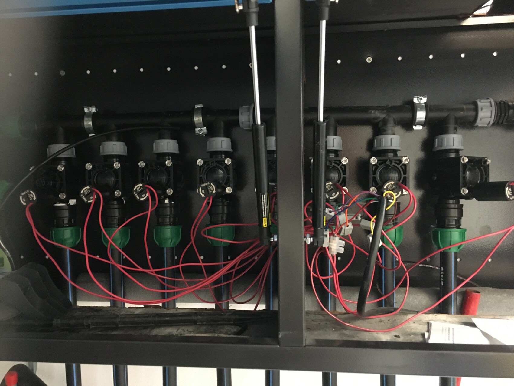 Mehrfachventil-System mit roten Kabeln, grünen Ventilköpfen und Rohrleitungen in einem Gehäuse.