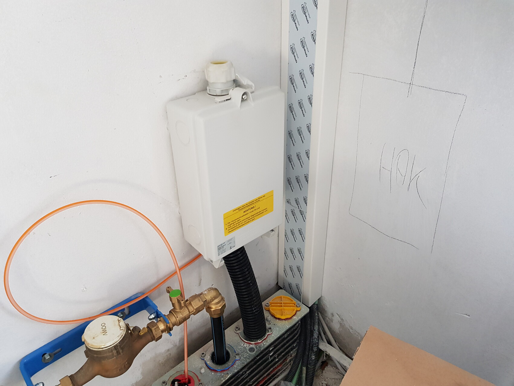 Weiße Installationsbox an der Wand, orange Schlauch, Messingventil und Kabel im Technikraum.