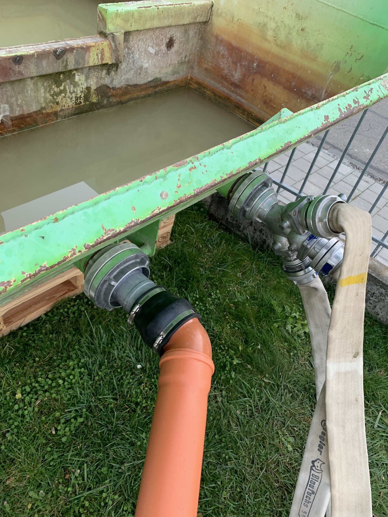 Grüne, rostige Metallwanne mit Wasser; unten orange Abflussrohr und Schlauchverbindungen.
