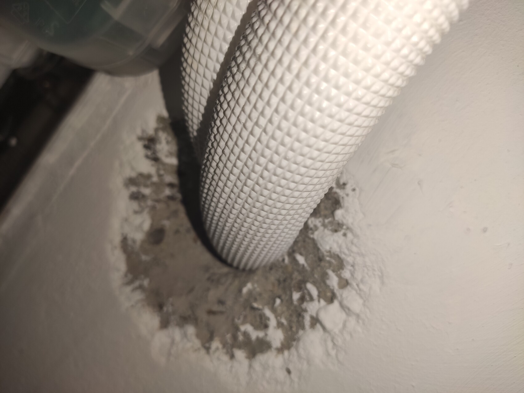 Weißer, geprägter flexibler Schlauch führt durch Loch in staubiger Wand mit Putzresten.