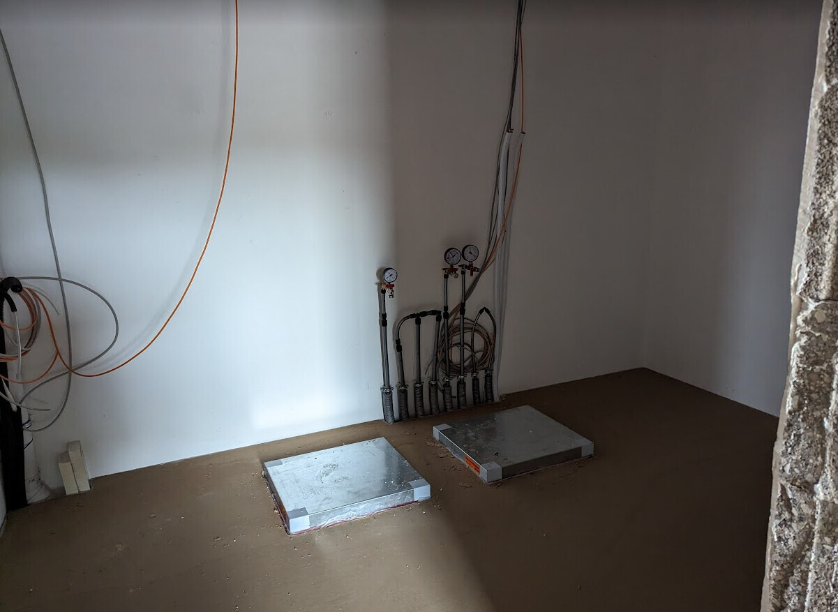Technikraum mit Rohren, Messgeräten, Kabeln; zwei Metallplatten auf dem Boden.