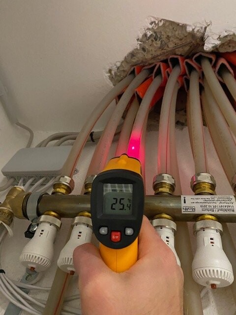 Infrarot-Thermometer wird von Hand vor Heizungsrohren gehalten; Anzeige 25,4 °C