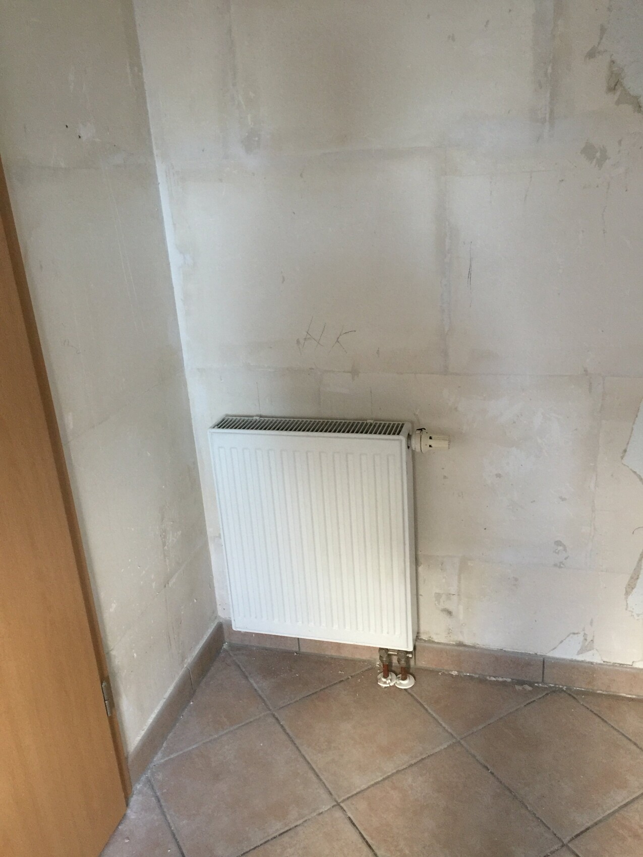 Weißer Heizkörper an einer unrenovierten Wand mit sichtbar installierten Rohren und Fliesenboden.