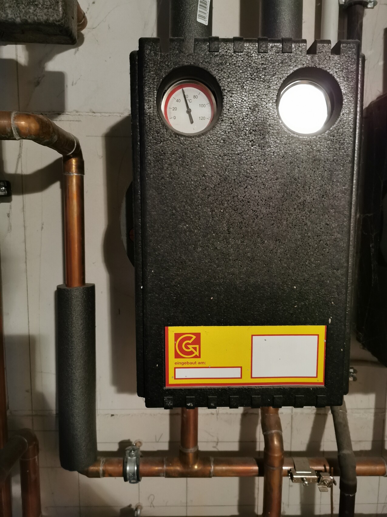Schwarzer Heizkessel an der Wand mit Temperaturanzeige, Kupferrohren und Gelb-Logo unten.
