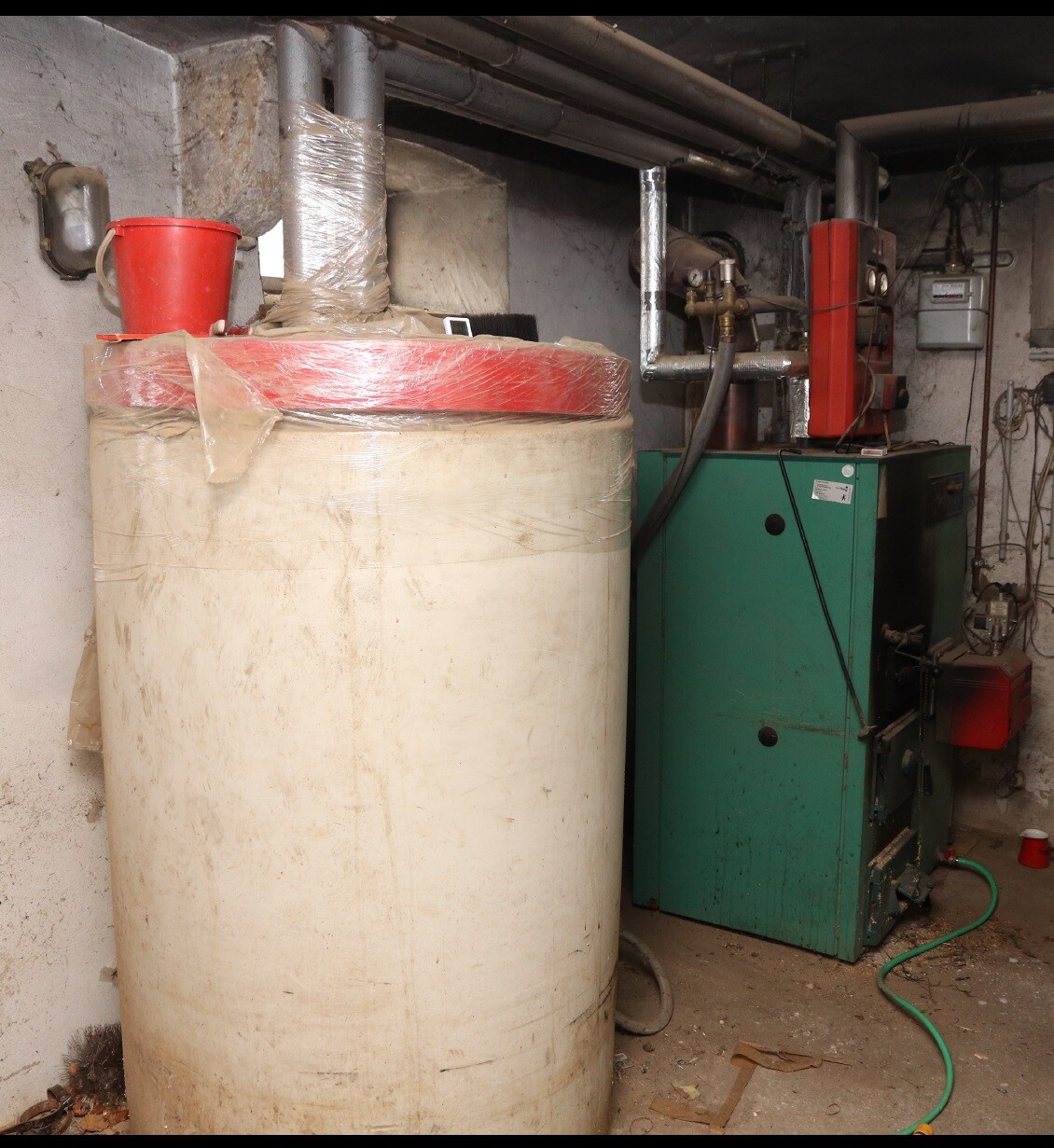 Großer Tank im Keller neben einer Heizungsanlage; Rohre, Schlauch und roter Eimer darauf.