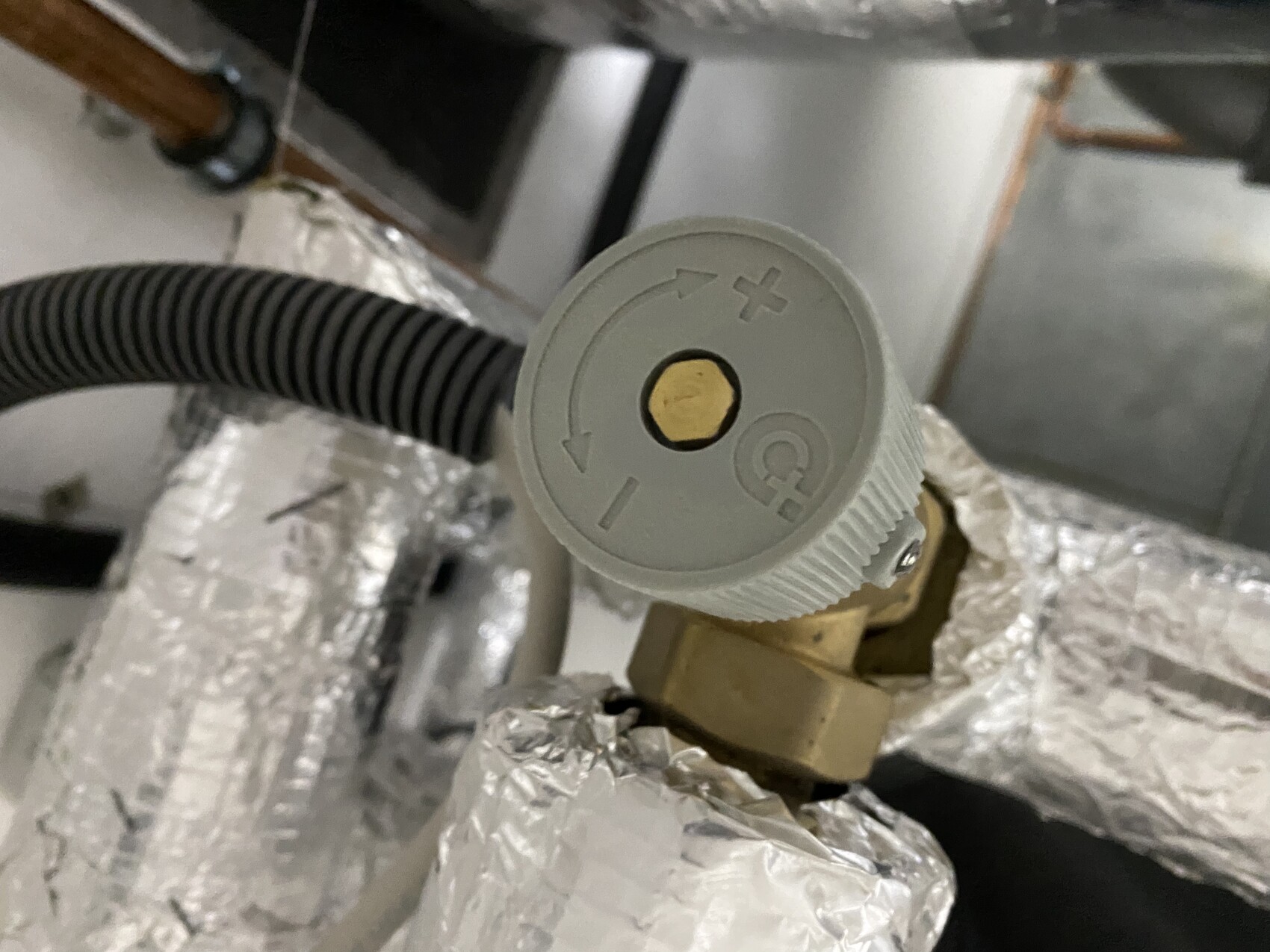 Grauer runder Ventilknopf mit Pfeilen, Pluszeichen und goldenem Schraubenkopf in der Mitte vor Rohren
