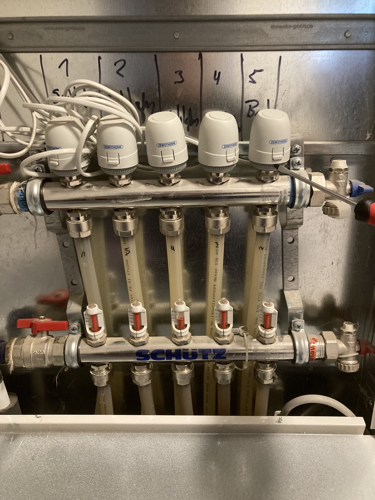 Verteiler mit sechs Rohren, roten Ventilen, weißen Thermo-Köpfen (ZEWOTHERM) und SCHÜTZ-Rohr.