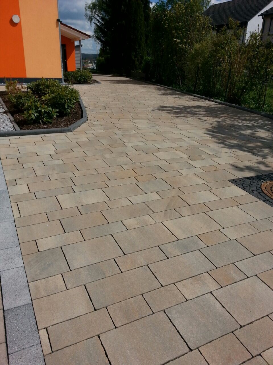 Heller Pflasterstein-Vorplatz und Einfahrt neben orangefarbener Hausfront mit Büschen