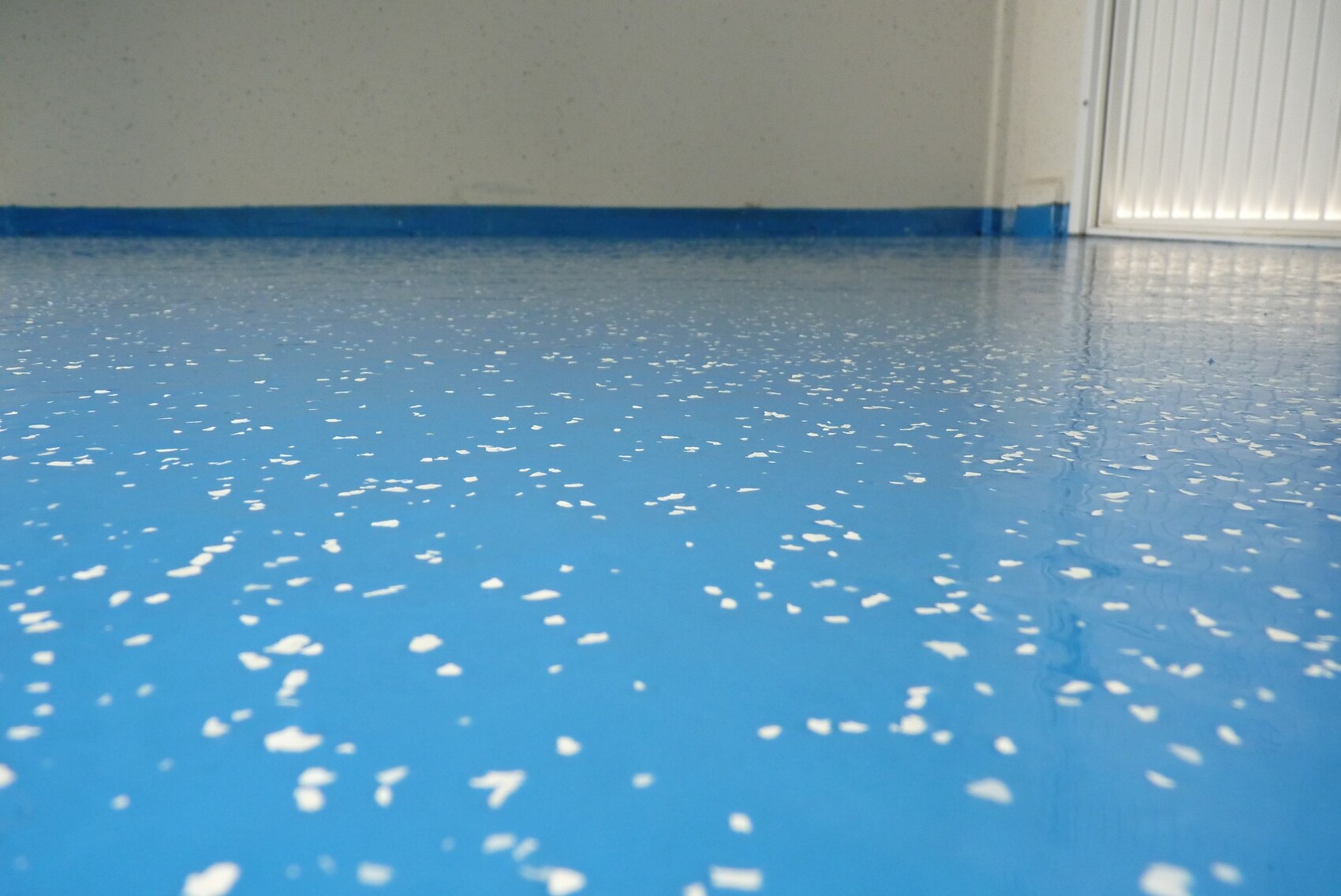 Blick auf glänzenden blauen Epoxidboden mit weißen Streuschips, weiße Wand und Tür rechts.
