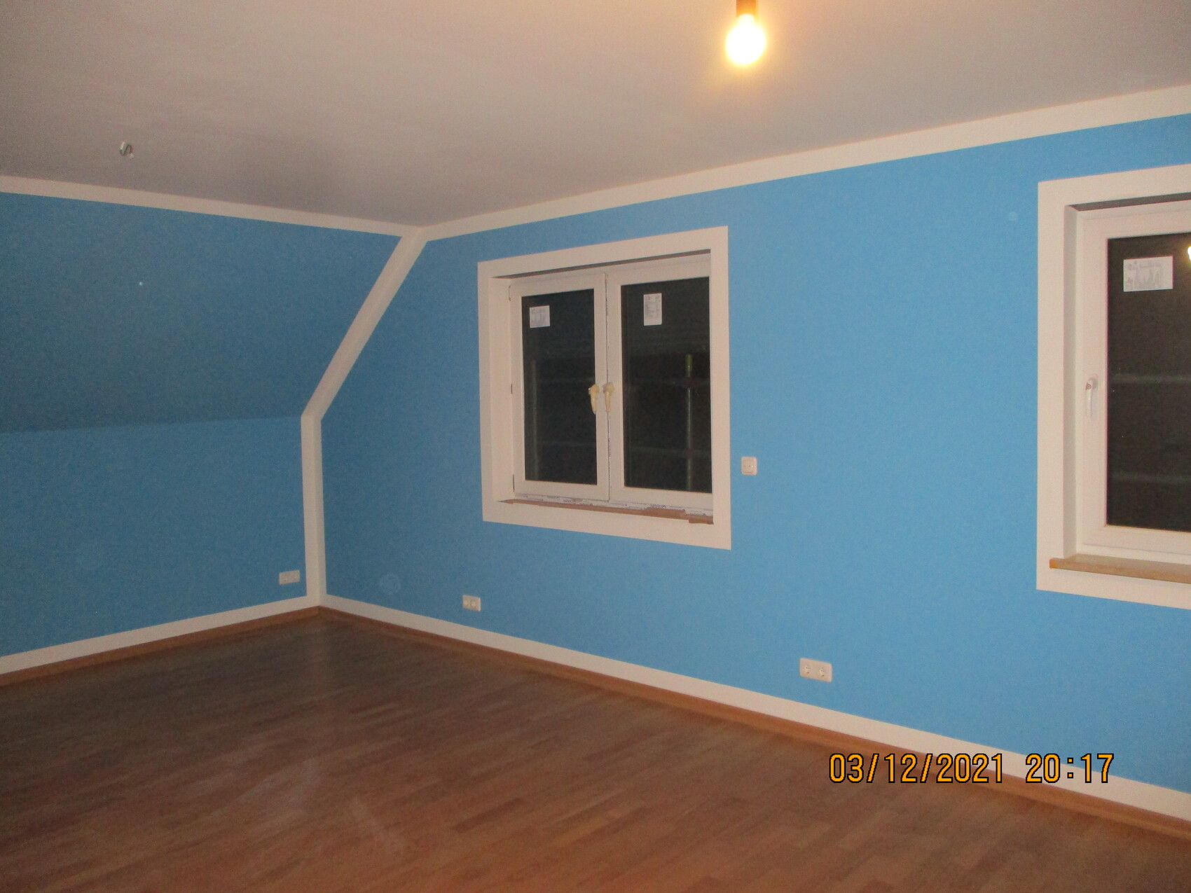 Blaue Zimmerwände mit weißem Rahmen, zwei Fenster, Holzboden und freihängende Glühbirne.
