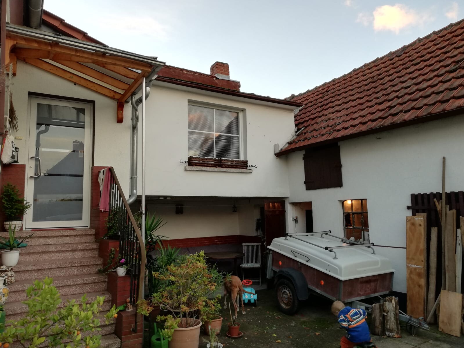 Hinterhof eines Hauses mit weißer Fassade, rotem Dach, Treppen und Pflanzen, Anhänger im Hof.