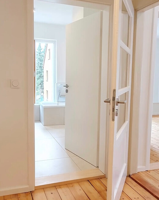 Offene Tür im Flur führt zu hellem Badezimmer mit weißer Badewanne am Fenster.