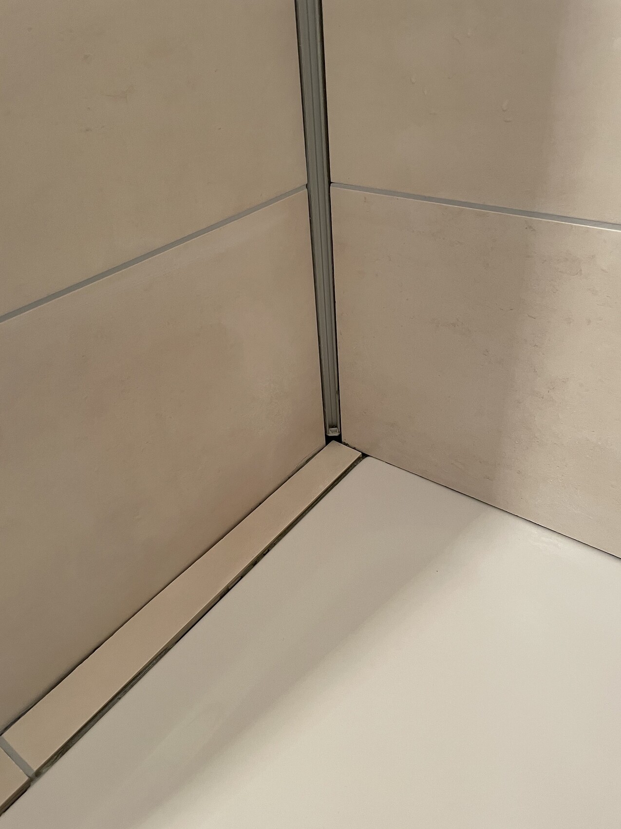 Eckdusche mit beigen Fliesen, Metallprofil in der Ecke und weißem Duschboden.