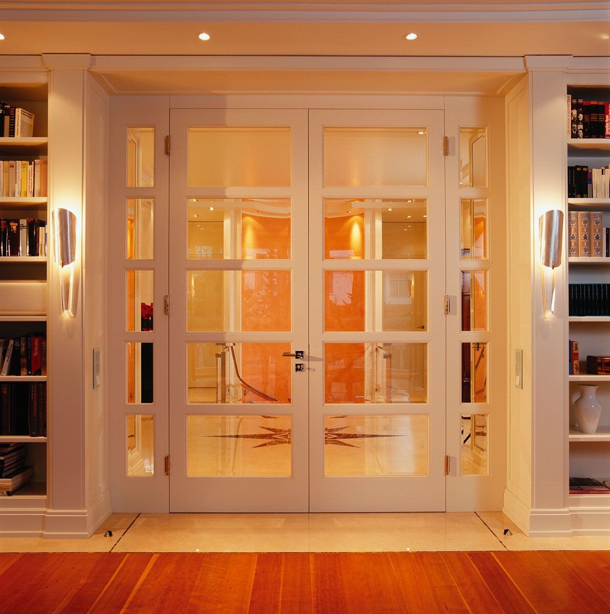 Weiße Doppeltüren mit Glasfenstern, flankiert von Bücherregalen, warm beleuchtet.