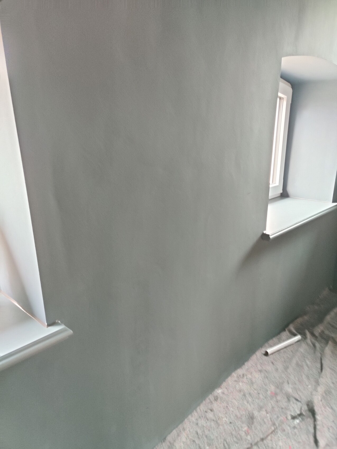 Innenraum mit grauer Wand, zwei Fensternischen und staubigem Boden bei Renovierungsarbeiten.