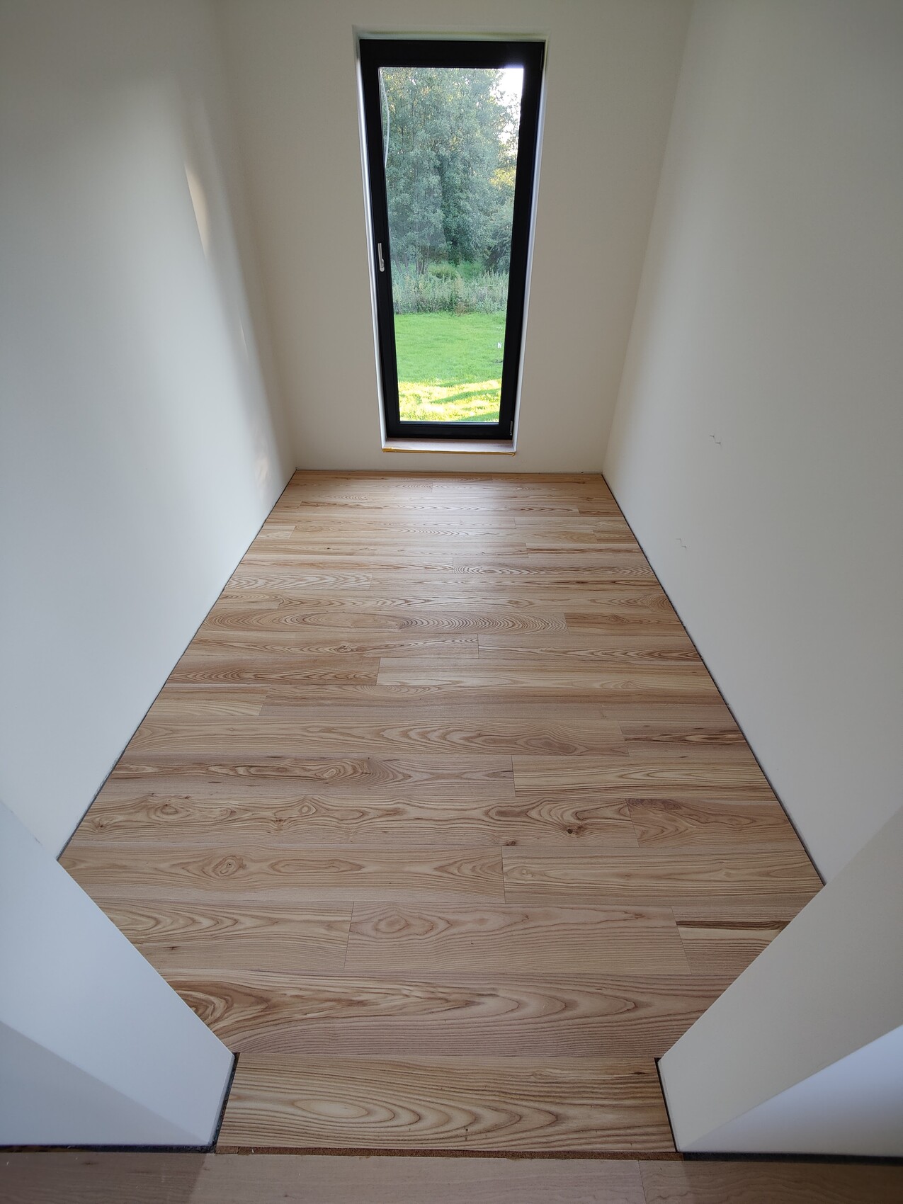 Kleiner heller Raum mit hellem Holzboden und schmalem vertikalem Fenster zum Garten.