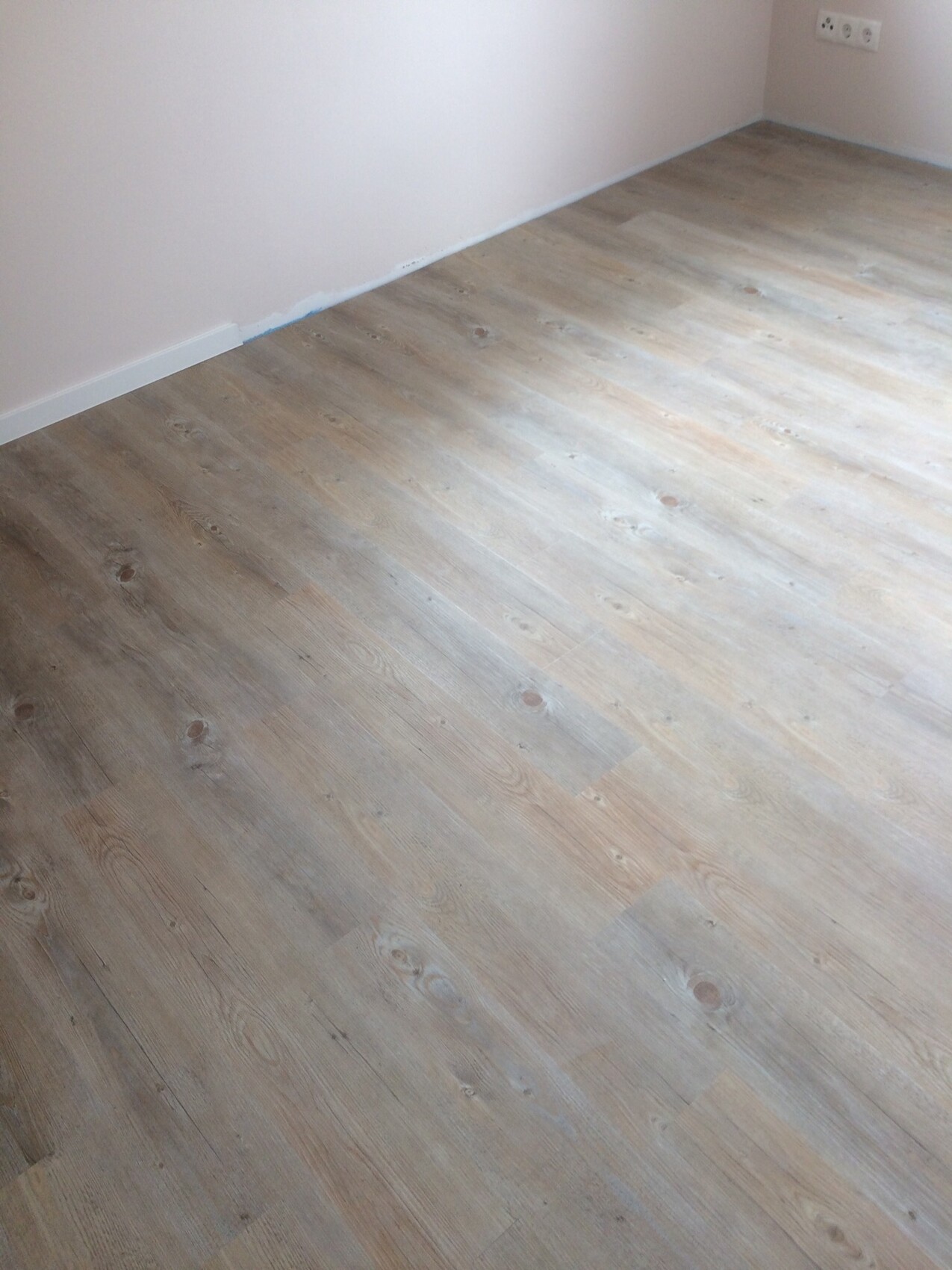 Helles, leeres Zimmer mit hellen Laminatdielen, rosa Wänden und Steckdose in der Ecke.
