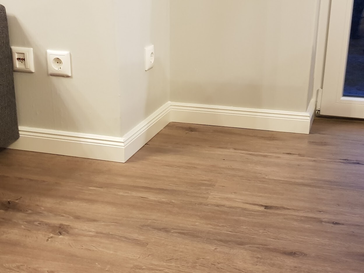 Heller Innenraum mit Holzboden, weißen Sockelleisten, Eckwand mit Steckdosen, Glastür rechts.