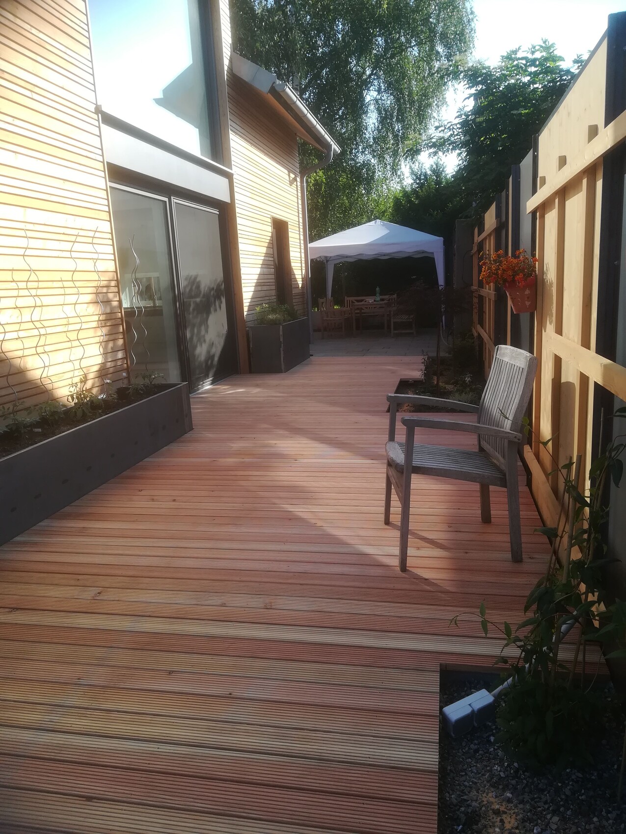 Holzdeck vor modernem Holzhaus mit Gartenmöbeln; Pavillon im Hintergrund, rechter Zaun.
